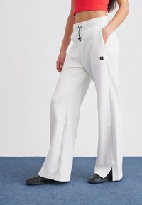 Ανοιχτό γκρι φόρμες με άνετη γραμμή, ρυθμιζόμενη μέση με κορδόνι, πλαϊνές τσέπες και μικρό λογότυπο μπροστά. Μαύρα παπούτσια slip-on.