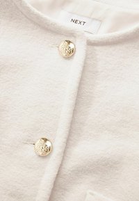 Cremfarbener Cardigan mit weicher Textur, ausgestattet mit zwei dekorativen goldfarbenen Knöpfen und einem runden Ausschnitt. Etikett: NEXT.