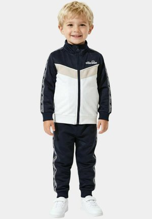 Jeune enfant portant un survêtement bleu marine et blanc avec fermeture éclair, des bandes avec logo sur les manches et les jambes, souriant et debout sur un fond blanc.