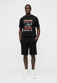 Άνδρας που φοράει μαύρο μπλουζάκι με γραφικό χαρακτήρα Peanuts, μαύρο σορτς, λευκές κάλτσες και λευκά αθλητικά παπούτσια, στέκεται με τα χέρια στις τσέπες.