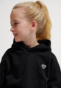 Schwarzer Kapuzenpullover mit einer Vordertasche, großer Kapuze und kleinem gesticktem Logo auf der Brust. Das Model hat blonde Haare, die zu einem Pferdeschwanz gebunden sind.