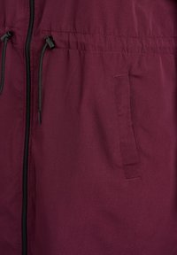 Kurtka w kolorze bordowym z zapięciem na suwak, regulowanymi czarnymi sznurkami i boczną kieszenią. Gładka tekstura i lekka tkanina.