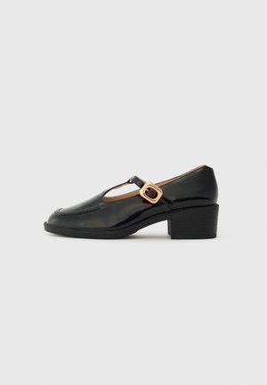 Black patent leather Mary Jane shoe with low block heel and gold buckle strap, side view on white background.