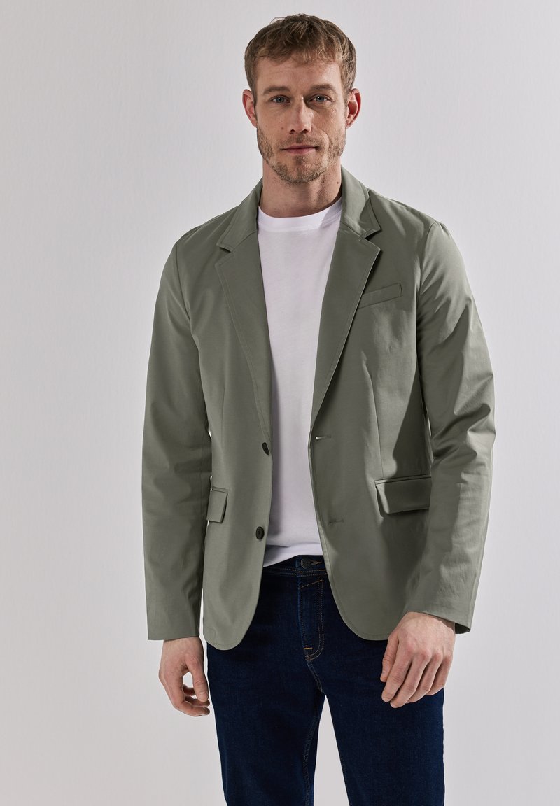 Blazer de mezcla de algodón en verde claro, con cierre de dos botones, solapas con notches y bolsillos laterales, llevado sobre una camiseta blanca y jeans oscuros.