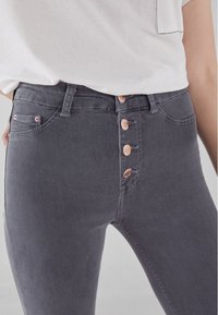 Mujer con pantalones ajustados de cintura alta grises, con cuatro botones de color rosa dorado visibles, y una camisa blanca metida por dentro.