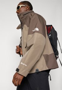 Mężczyzna w beżowej i brązowej kurtce Gore-Tex North Face oraz czarnej torbie na plecy, trzymający pasek, w białych okularach przeciwsłonecznych, zwrócony w lewo.