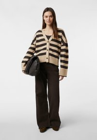 Sweter w paski w kolorze beżowym i ciemnobrązowym, z dekoltem w serek, guzikami i szerokimi rękawami, zestawiony z ciemnobrązowymi spodniami z wysokim stanem i czarną torbą.