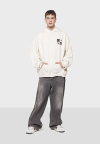 Sudadera beige de gran tamaño con un bolsillo frontal y cuadrados gráficos en negro. Combinada con jeans de denim gris sueltos y zapatillas blancas.