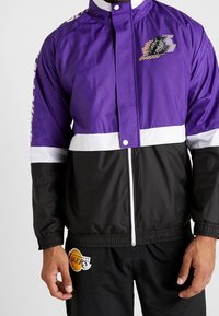 Lila och svart vindjacka med ett vitt rand, med en logotyp och grafiska detaljer. Tillverkad av ett mjukt, lätt tyg. Dragkedjeslutning.