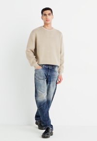 Beige long-sleeve sweater, light texture; blue denim jeans with a faded finish; black shoes. Standing pose against a plain background.