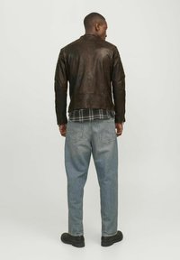 Braune Lederjacke mit taillierter Passform und strukturiertem Oberflächenmaterial; getragen über einem karierten Hemd und hellblauen lässig geschnittenen Jeans. Schwarze Schuhe.
