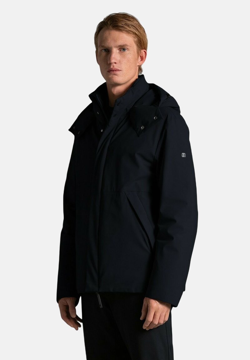 Junger Mann mit kurzen, rötlichen Haaren, der eine schwarze, gefütterte Winterjacke mit Kapuze trägt und vor einem einfarbigen, hellen Hintergrund steht.