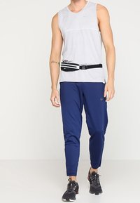 Top atletico grigio senza maniche, pantaloni tapered blu navy, sneakers nere e una cintura da corsa nera con accenti bianchi. Tessuto liscio, design aderente.