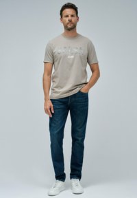 Hombre que lleva una camiseta taupe de "SALSA JEANS", jeans oscuros y zapatillas blancas, de pie con una mano en el bolsillo contra un fondo liso.
