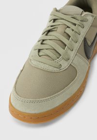 Artimas smūgis įrengtas smėlio spalvos sportbačiui su žaliais raišteliais, zomšos ir tinklinio viršumi, juodu logotipu ir gumine guma padėtyje ant balto fono.