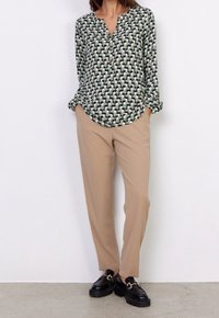 Blusa a maniche lunghe con un motivo geometrico verde e crema, abbinata a pantaloni beige a sigaretta. Mocassini neri con dettagli in metallo dorato.
