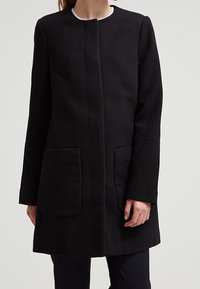 Černý vlněný kabát s minimalistickým designem, který má kulatý výstřih, dlouhé rukávy a dvě přední kapsy pro funkčnost.