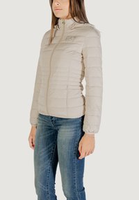 Giacca puffer beige chiaro con colletto alto, chiusura con zip e dettagli di cucitura orizzontale. Caratterizzata da polsini elasticizzati e un logo sul petto.