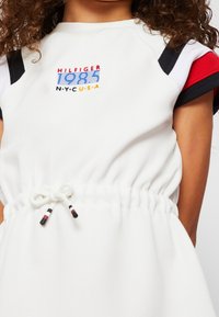 Weißes Kleid mit kurzen Ärmeln, das mit roten, marineblauen und weißen Akzenten verziert ist. Mit Zugband in der Taille und gesticktem "HILFIGER 1985 N.Y.C.U.S.A." Detail.