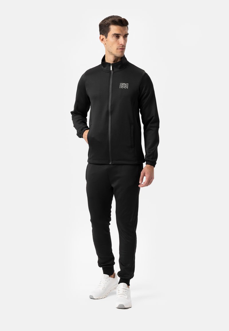 Zwart atletisch trainingspak met een jas met ritssluiting en hoge kraag, zijzakken, aansluitende manchetten, en bijpassende trainingsbroek, gecombineerd met witte sneakers.