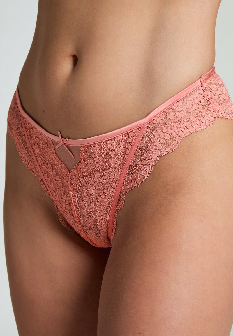 Kant bikini slip in zacht perzik, met ingewikkelde bloemenpatronen, geschulpte randen en een decoratieve strik aan de voorkant.