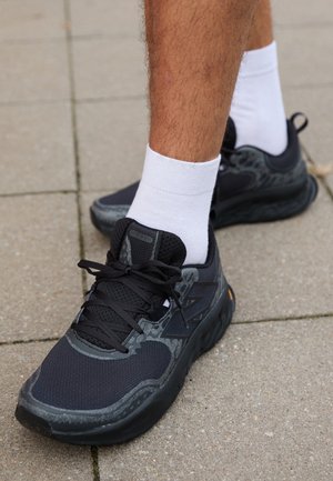 Personne portant des chaussures de sport noires et grises avec des chaussettes blanches, debout sur un trottoir carrelé en plein air.