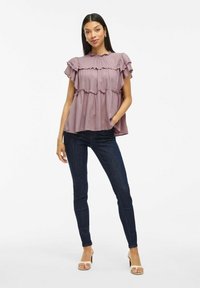 Top viola con maniche a ruffle e design a strati, abbinato a jeans blu scuro e sandali con tacco color marrone chiaro. Tessuto dalla superficie liscia.