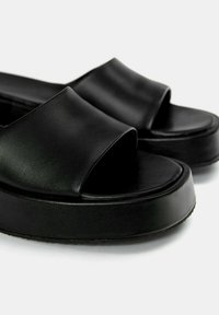 Black slide sandals feature a smooth leather upper, wide strap, and thick platform sole. The design is minimalist and modern.