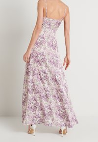 Robe maxi fleurie à fines bretelles, présentant un motif lilas et vert sur un fond pâle, tissu fluide et ourlet légèrement évasé.