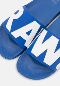 Blue slide sandals with wide straps featuring bold white lettering. Textured footbed provides grip. Smooth upper material in a shiny finish.