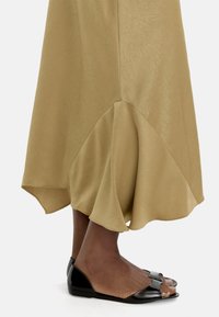 Oliwkowa spódnica z rozkloszowanym dołem i delikatną teksturą, zestawiona z czarnymi sandałami na otwartym palcu o gładkim wykończeniu i minimalistycznym designie.