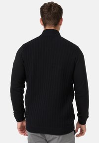 Gilet en tricot noir avec un motif texturé et des manches longues noires unies. Dispose d'un col haut et de poignets côtelés pour un ajustement structuré.