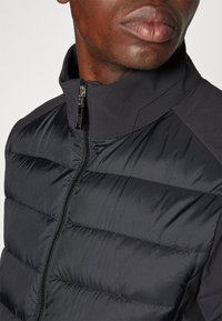 Gros plan sur une personne portant une veste matelassée noire avec un col montant et une fermeture éclair argentée partiellement ouverte.