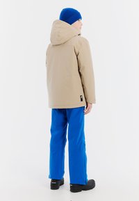 Beige capuchonjack met een gladde textuur, gecombineerd met blauwe waterdichte broek en zwarte laarzen, gezien van achteren tegen een witte achtergrond.