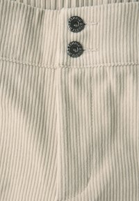 Close-up van crème corduroy stof met twee zwarte knopen met het label "STUDIO" op een taille-naad.