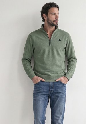 Mann mit dunklen Haaren und Bart, trägt grünes Viertel-Zip-Sweatshirt und blaue Jeans, Hände in den Taschen, schaut nach rechts an einer einfarbigen Wand.