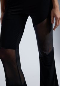Leggings noirs avec des inserts en panneaux transparents. Le matériau est lisse avec un design ajusté, mettant en valeur la forme des jambes et les contrastes de texture.