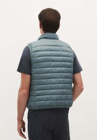 Lichtblauwe, gewatteerde gilet met een hoge kraag, met een gladde textuur en horizontale stiksel details, gedragen over een donker overhemd.