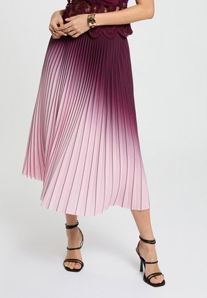 Donna che indossa una gonna midi plissettata con sfumatura ombré dal viola al rosa, abbinata a sandali neri con cinturini e tacco alto.