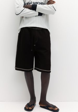 Persona che indossa pantaloncini neri fino al ginocchio, camicia bianca a maniche lunghe con scritta e infradito nere, in piedi con le braccia incrociate.