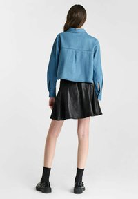 Korte blauwe denimjas met lange mouwen, een rugplooi en open manchetten, gecombineerd met een zwarte leren rok met uitlopende onderkant en zwarte schoenen.