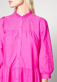 Robe en coton rose avec encolure à volants, boutonnage sur le devant et broderies sur les manches, avec un design fluide et froncé.