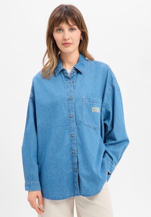 Junge Frau mit hellbraunen Haaren trägt ein übergroßes blaues Jeanshemd mit "American Vintage" auf der Brusttasche und beige Hose.