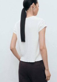 Bílá krátká halenka s mírně zkráceným lemem, měkkou texturou a jednoduchým designem, kterou doplňují tmavě hnědé kalhoty.