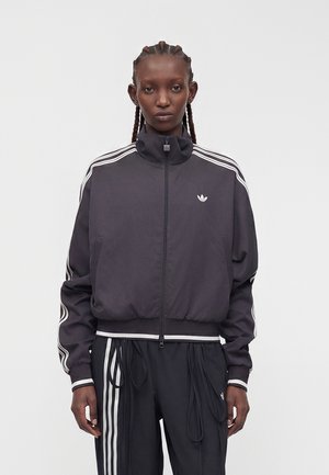 Persona con capelli intrecciati che indossa una giacca e pantaloni sportivi Adidas neri con strisce bianche, in piedi davanti a uno sfondo chiaro e uniforme.