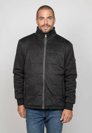 Homme portant une veste matelassée noire avec des manches texturées et un jean bleu, debout devant un fond gris uni.