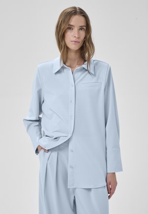 Mujer con camisa suelta de botones azul claro y pantalones plisados a juego, de pie contra un fondo gris liso con una mano en el bolsillo.