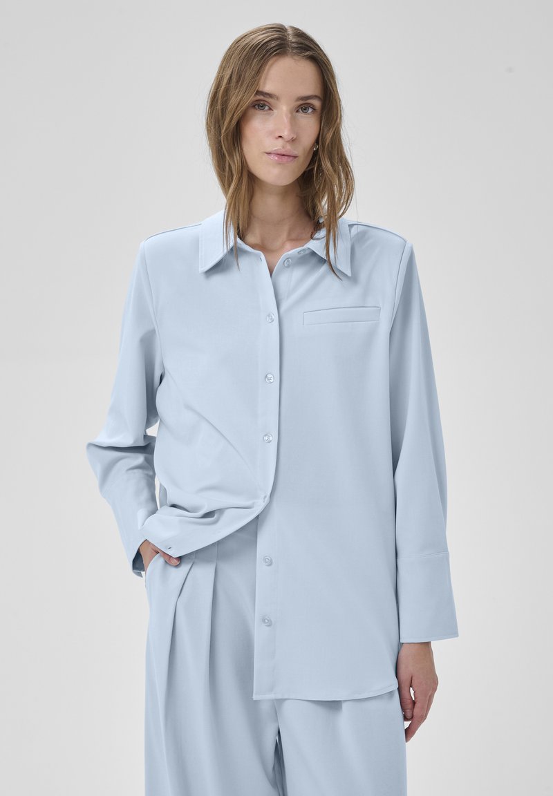 Žena oblečená v svetlomodrej voľnej košeli so zapínaním na gombíky a v rovnakých plisovaných nohaviciach, stojaca na jednoduchom sivom pozadí s jednou rukou v vrecku.