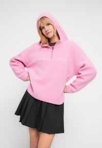 Νεαρή γυναίκα με ξανθά μαλλιά, που φορεί ροζ φλις φούτερ με κουκούλα και μαύρη πλισέ φούστα, ποζάρει με τα χέρια στη μέση μπροστά σε λευκό φόντο.