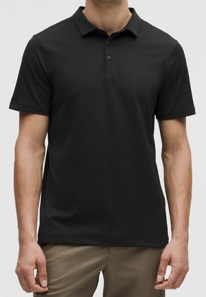 Mann trägt ein schlichtes schwarzes Kurzarm-Poloshirt mit drei Knöpfen, kombiniert mit hellbraunen Hosen, gezeigt von Hals bis Hüfte.
